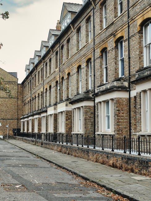 A quiet residential street featuring a row of Victorian-style terraced houses with white facades, decorative bay windows, and red-tiled roofs, located on a council estate in Tooting. The street has a 