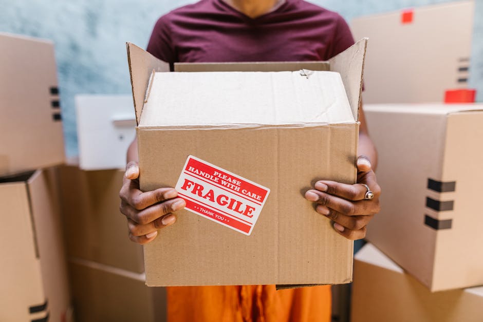 Close-up of a person holding an open cardboard box with both hands, revealing its empty interior. The individual is standing indoors among other sealed cardboard boxes, some marked with black tape, indicating a packing and moving process. The person's hands are visible, with a silver ring on one finger, and the background includes additional boxes stacked against a light-colored wall, suggesting a home or storage environment prepared for a house relocation. The box features a red and white 'FRAGILE' sticker, emphasizing careful handling during the moving service provided by Tooting Removals. The scene illustrates the packing and loading phase of a household move, highlighting the importance of proper packaging materials such as cardboard boxes and labeling to ensure safe furniture transport or home relocation, aligned with the Tooting Broadway removals guide for local moves.
