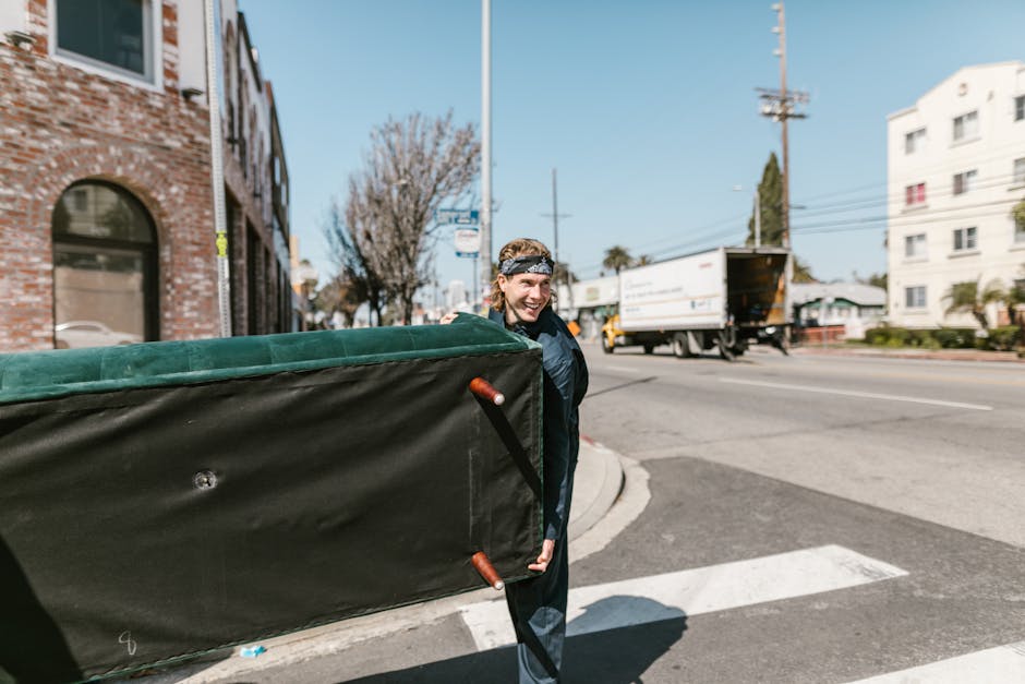 A young man with long hair wearing a headband and dark clothing is smiling and carrying a large, padded furniture blanket with orange handles, preparing for a home relocation. He is standing on a sidewalk at an urban street corner next to a black trolley or dolly, which is supporting the furniture blanket. In the background, a white delivery truck with its rear door open is parked on the street, ready for loading or unloading furniture and packed boxes, some of which are wrapped in cardboard and plastic for protection during the moving process. The scene includes a brick building with arched windows on one side and a white apartment building on the other, with a clear blue sky overhead and leafless trees indicating a cooler season. The surroundings suggest an organized furniture transport or packing and moving activity by Tooting Removals, as part of their house removals services in the area.