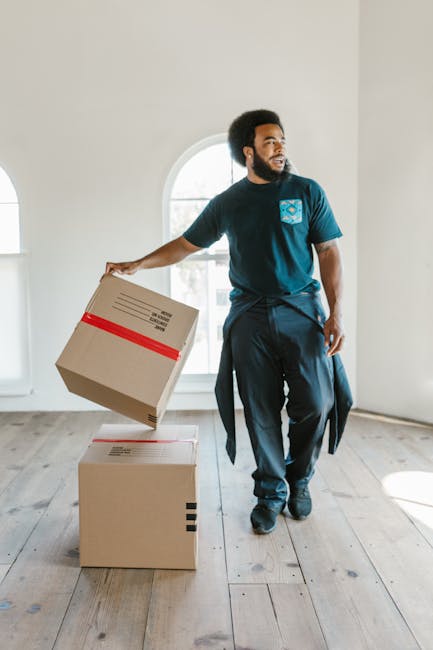 Close-up of a person holding an open cardboard box with both hands, revealing its empty interior. The individual is standing indoors among other sealed cardboard boxes, some marked with black tape, indicating a packing and moving process. The person's hands are visible, with a silver ring on one finger, and the background includes additional boxes stacked against a light-colored wall, suggesting a home or storage environment prepared for a house relocation. The box features a red and white 'FRAGILE' sticker, emphasizing careful handling during the moving service provided by Tooting Removals. The scene illustrates the packing and loading phase of a household move, highlighting the importance of proper packaging materials such as cardboard boxes and labeling to ensure safe furniture transport or home relocation, aligned with the Tooting Broadway removals guide for local moves.